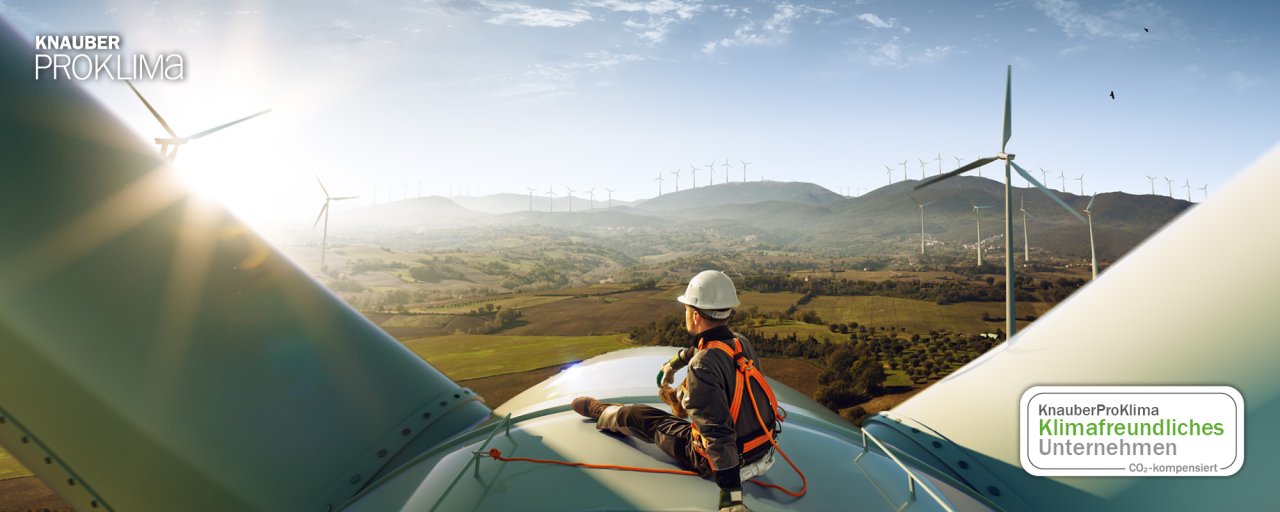 Arbeiter mit Helm und Schutzkleidung sitzt auf Windradflügel mit Blick auf Windpark und Landschaft bei klarem Himmel.