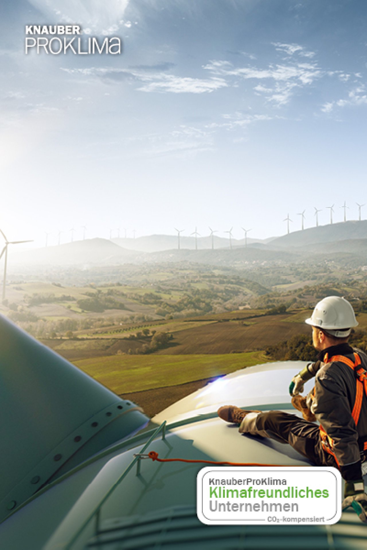 Techniker mit Schutzhelm und Sicherheitsausrüstung sitzt auf Windkraftanlage vor Landschaft mit Windrädern bei Sonnenaufgang.