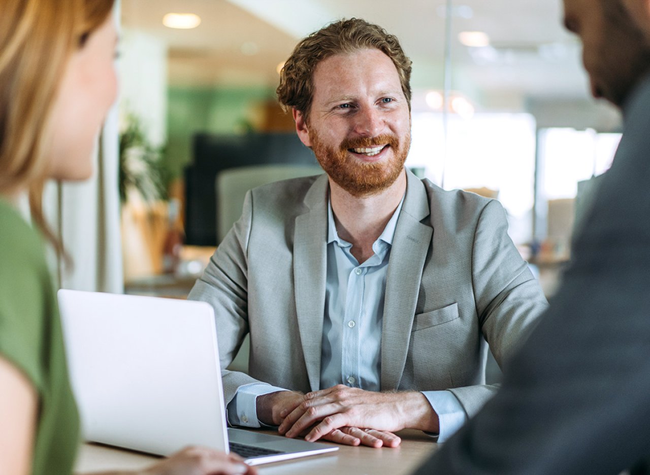Ein bärtiger Mann sitzt an einem Schreibtisch und lacht ein Paar an.