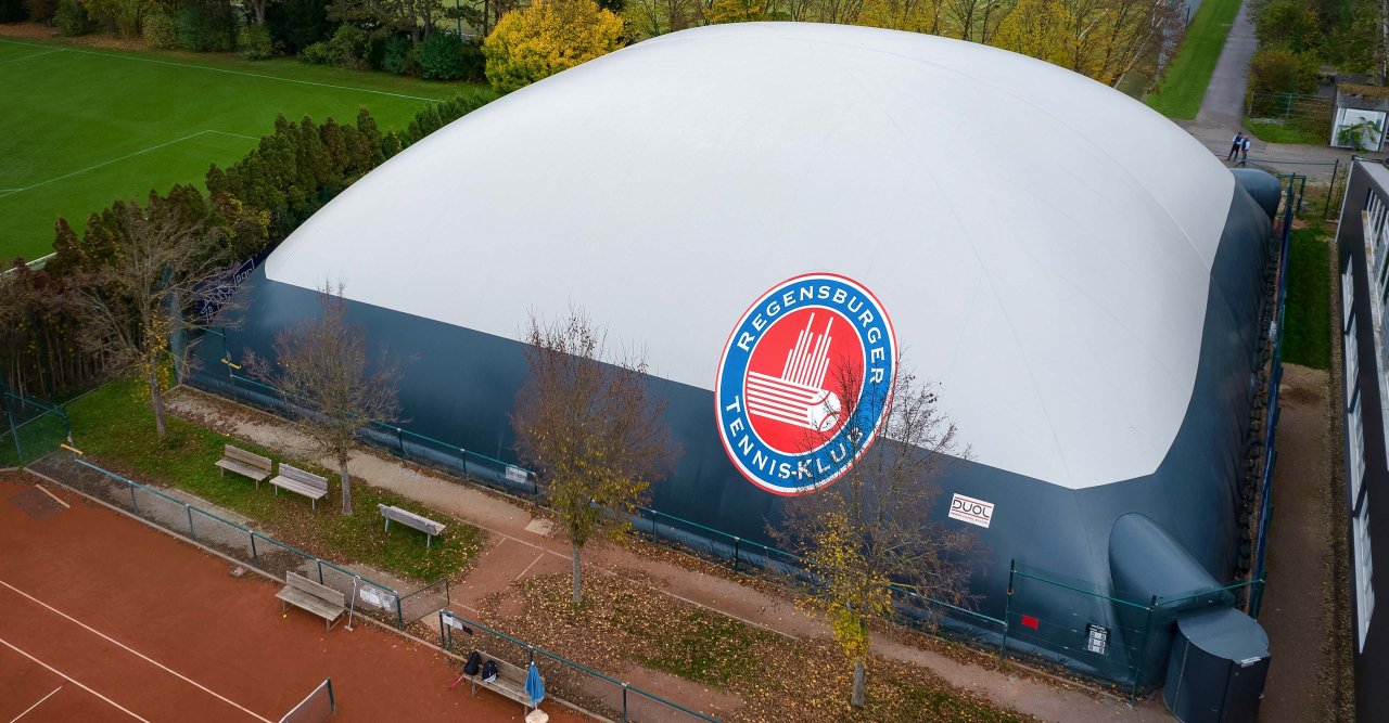 Große weiße Tennis-Halle mit blauem Sockel und Logo des Regensburger Tennis-Klubs, umgeben von Tennisplätzen und Bäumen.