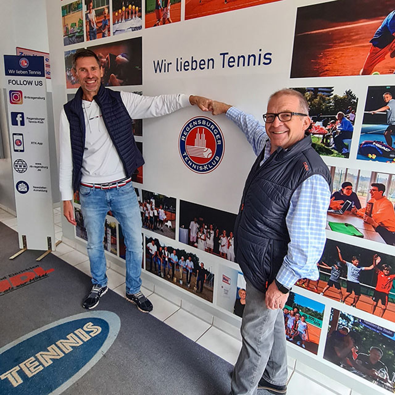 Zwei Männer in Freizeitkleidung geben sich vor einer Wand mit Tennisfotos und dem Schriftzug 'Wir lieben Tennis' einen Faustgruß.