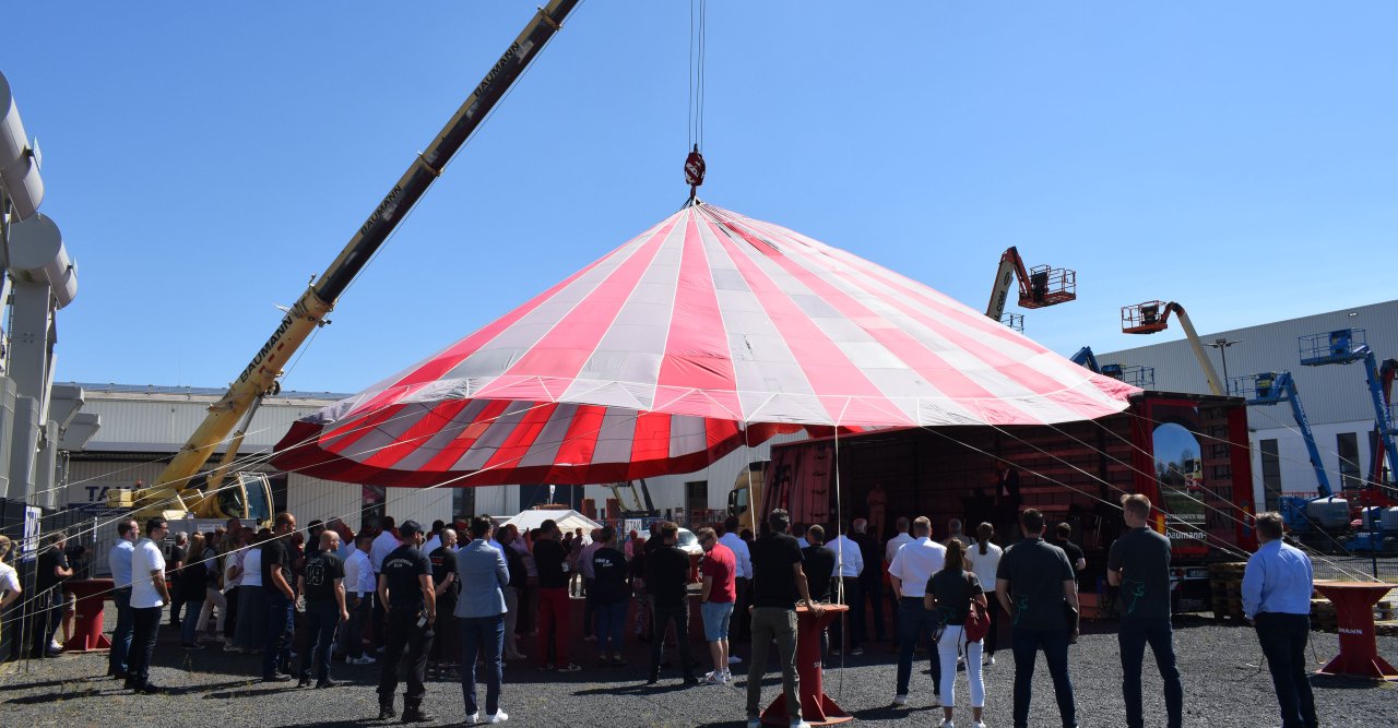 Ein Platz voller Menschen, in der Mitte hängt ein rot-weißes Zeltdach von einem Kran.