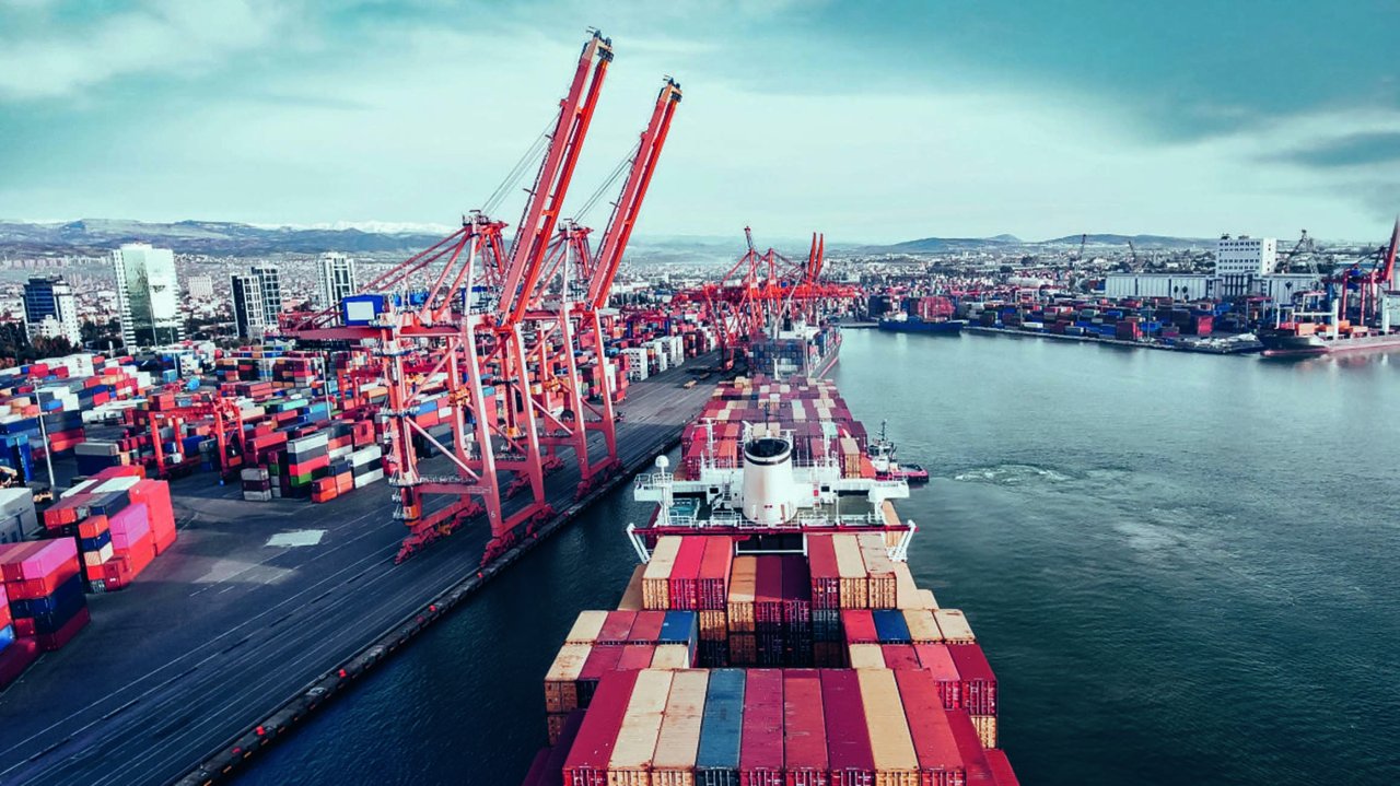 Containerschiff beladen mit bunten Containern im Hafen, umgeben von großen Hafenkränen und Lagerflächen.