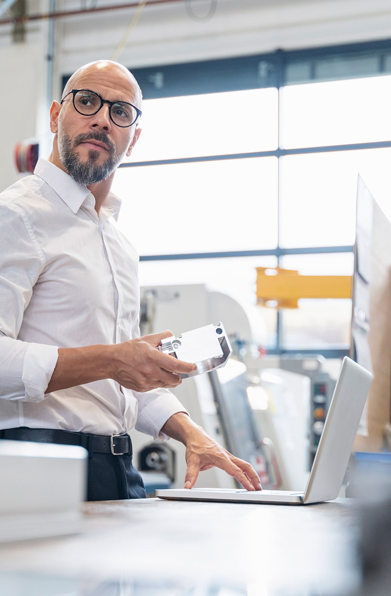 Mann mit Brille und weißem Hemd hält ein Smartphone und arbeitet an einem Laptop in einem hellen Büro.
