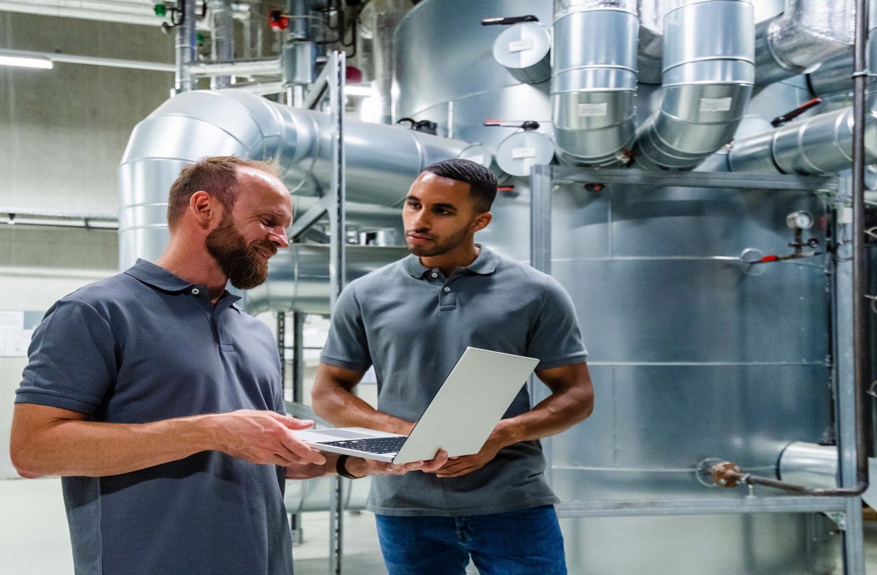 Zwei Techniker in grauen Poloshirts besprechen Daten an einem Laptop in einer Industrieanlage mit großen Metallrohren.