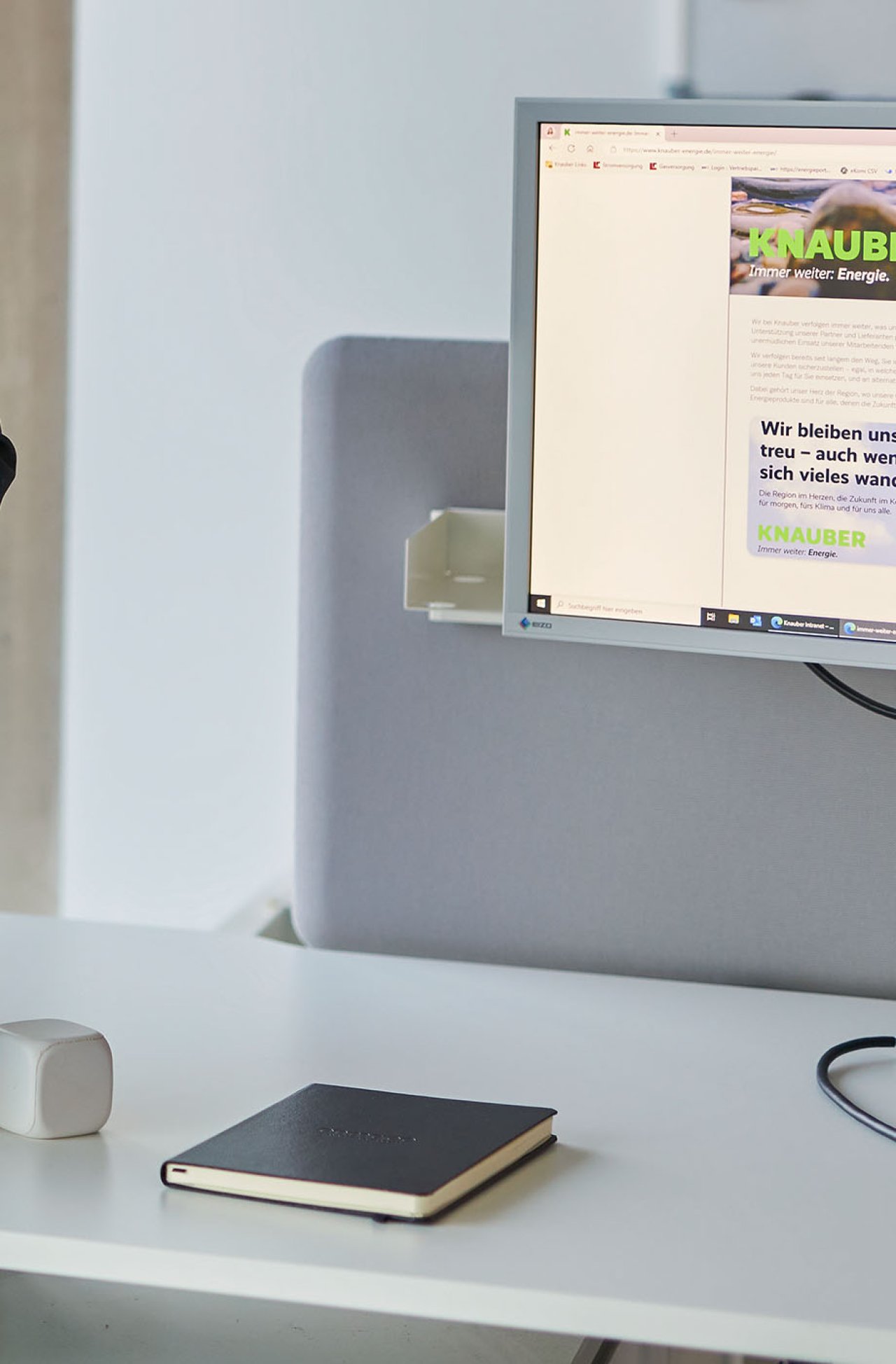 Ein Schreibtisch mit einem Notizbuch, im Hintergrund ist ein Bildschirm zu sehen.